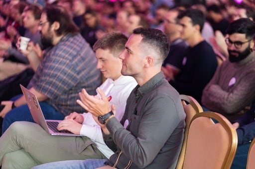 Two men in the audience, the one in the background is working on his laptop